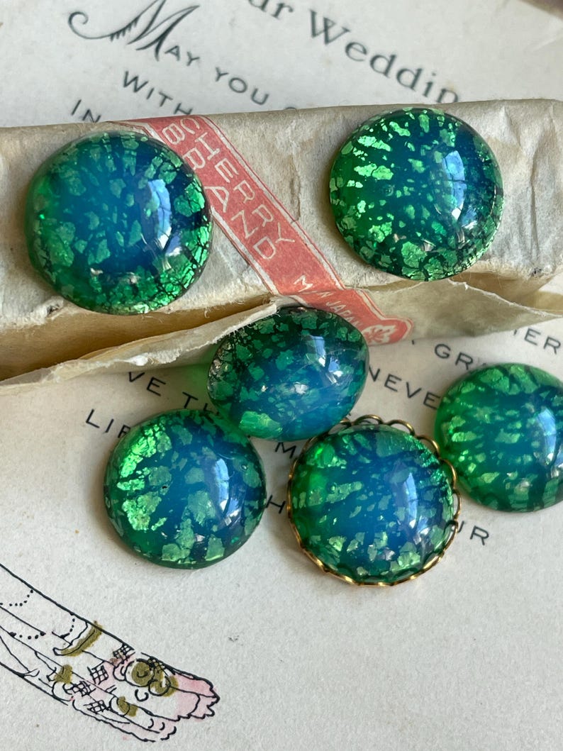 May include: Five round, iridescent green and blue glass cabochons with gold foil flecks.  Four of the cabochons are plain and one has a gold metal rim.