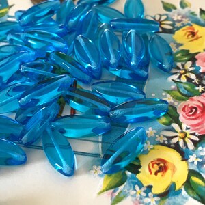 May include: A pile of turquoise blue glass beads, each bead is oval shaped and has a slightly frosted finish.