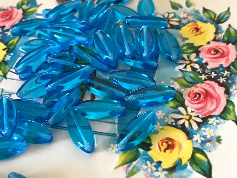 May include: A pile of turquoise blue glass beads, each bead is oval shaped and has a slightly frosted finish.