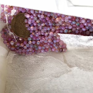 K&ouml;nnte beinhalten: Ein wei&szlig;er Papierbeutel gef&uuml;llt mit einer gro&szlig;en Menge kleiner, irisierender rosa und lila Perlen. Eine goldene M&uuml;nze ist in der Mitte des Bildes zu sehen.