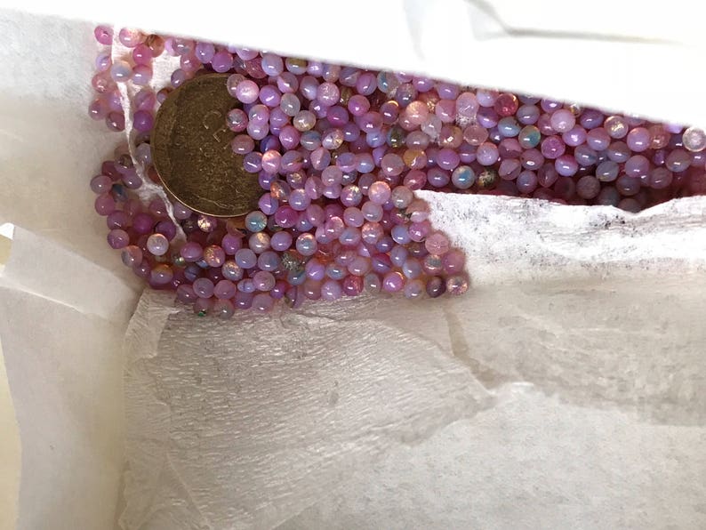 K&ouml;nnte beinhalten: Ein wei&szlig;er Papierbeutel gef&uuml;llt mit einer gro&szlig;en Menge kleiner, irisierender rosa und lila Perlen. Eine goldene M&uuml;nze ist in der Mitte des Bildes zu sehen.