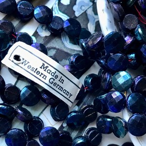 Puede incluir: Una hebra de cuentas de vidrio azul oscuro iridiscente con una etiqueta que dice "Made in Western Germany".