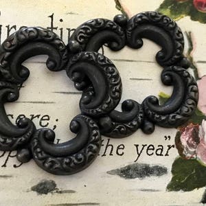 May include: Two ornate, dark gray decorative frames with scrollwork and a vintage aesthetic. The frames are set against a backdrop of faded text and floral illustrations, evoking a classic, antique style.