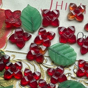 May include: Close-up of various red flower and green leaf-shaped glass beads. The flower beads have a faceted design, and the leaves have detailed veining. The beads are scattered on a decorative background.