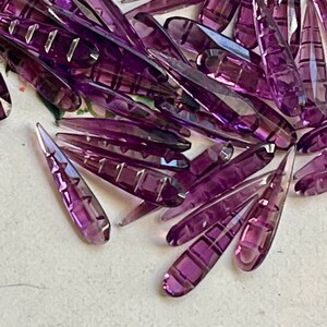 May include: A large pile of purple glass teardrop-shaped beads with a faceted design. The beads are arranged in a random pattern and are all the same size and shape.