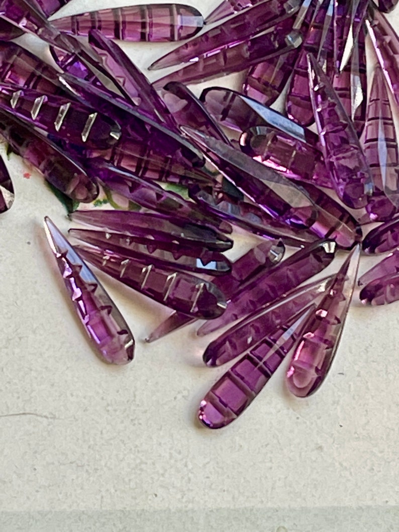 May include: A large pile of purple glass teardrop-shaped beads with a faceted design. The beads are arranged in a random pattern and are all the same size and shape.