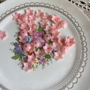 Peut inclure: Une assiette de boutons en plastique rose en forme de fleur. Les boutons sont dispos&eacute;s de mani&egrave;re al&eacute;atoire sur une assiette blanche avec un motif floral dor&eacute;.