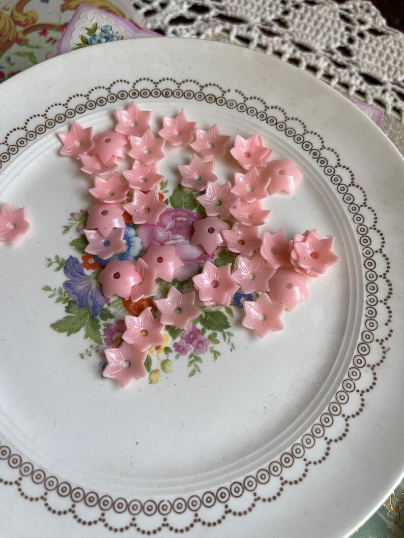 Peut inclure: Une assiette de boutons en plastique rose en forme de fleur. Les boutons sont dispos&eacute;s de mani&egrave;re al&eacute;atoire sur une assiette blanche avec un motif floral dor&eacute;.