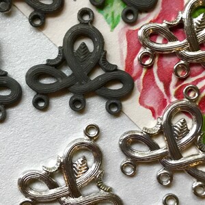 May include: A close-up of silver and black metal decorative embellishments with intricate designs. The embellishments have multiple loops and a leaf-like design in the center.