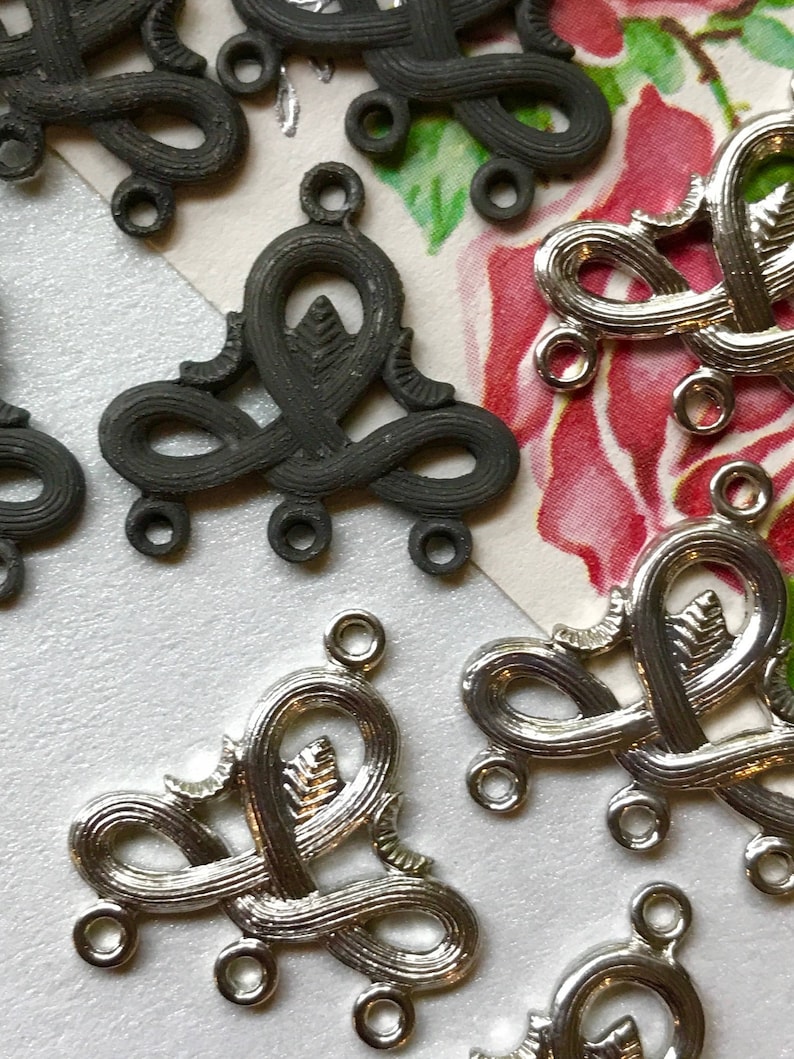 May include: A close-up of silver and black metal decorative embellishments with intricate designs. The embellishments have multiple loops and a leaf-like design in the center.