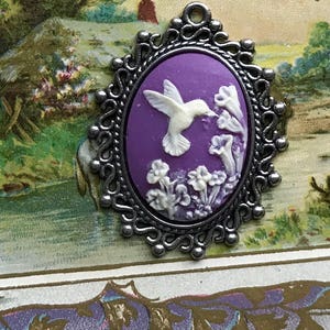May include: A silver cameo pendant with a purple background and a white hummingbird in flight. The hummingbird is depicted with its beak in a flower. The cameo is surrounded by a silver filigree frame.