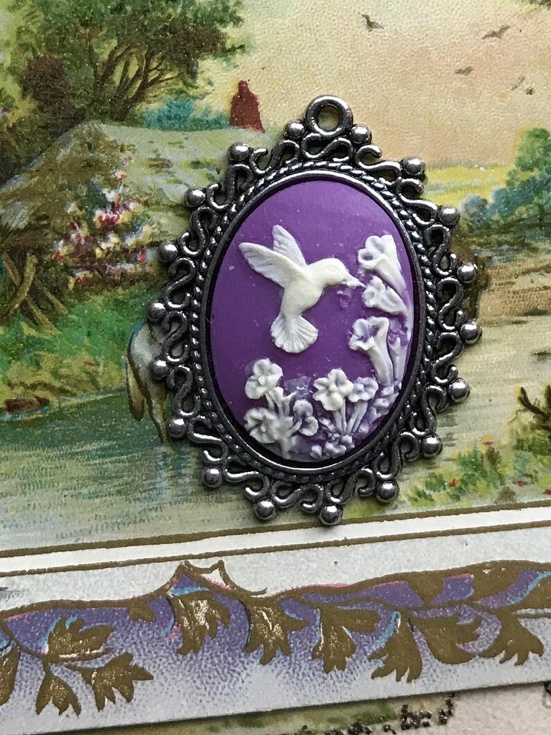 May include: A silver cameo pendant with a purple background and a white hummingbird in flight. The hummingbird is depicted with its beak in a flower. The cameo is surrounded by a silver filigree frame.