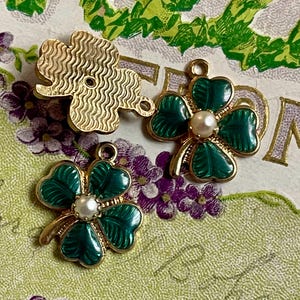 Puede incluir: Tres colgantes de tr&eacute;bol de oro y verde. Dos colgantes presentan hojas de tr&eacute;bol de esmalte verde con un centro de perla de imitaci&oacute;n. El tercer colgante es un tr&eacute;bol dorado texturizado. Los colgantes est&aacute;n sobre un fondo floral.