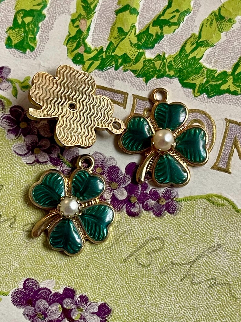 Puede incluir: Tres colgantes de tr&eacute;bol de oro y verde. Dos colgantes presentan hojas de tr&eacute;bol de esmalte verde con un centro de perla de imitaci&oacute;n. El tercer colgante es un tr&eacute;bol dorado texturizado. Los colgantes est&aacute;n sobre un fondo floral.