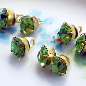 May include: Close-up of several green gemstone studs with gold-colored settings. The gemstones are faceted and have a bright, vibrant green color. The studs are arranged on a white surface with blue and green floral accents.