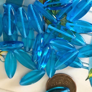 May include: A pile of turquoise blue glass beads in the shape of long oval leaves. The beads are translucent and have a slight shimmer.