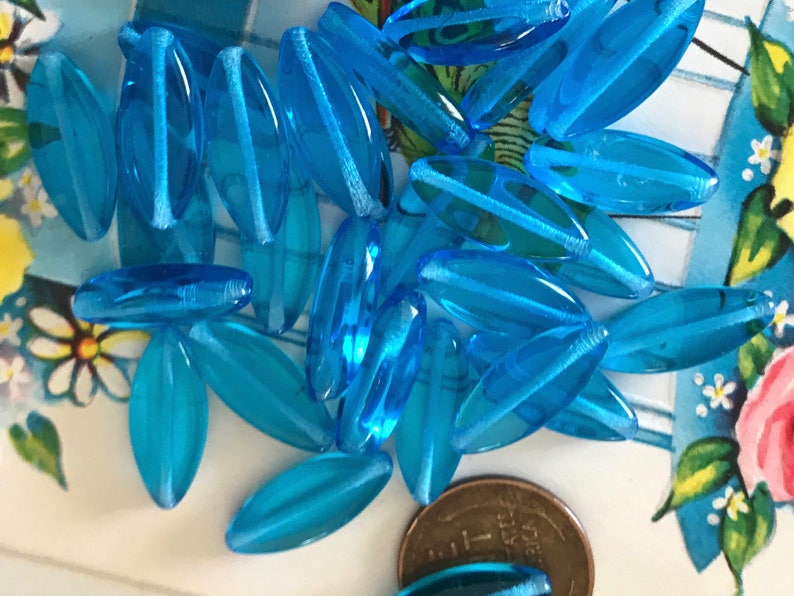 May include: A pile of turquoise blue glass beads in the shape of long oval leaves. The beads are translucent and have a slight shimmer.