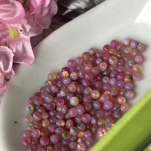 K&ouml;nnte beinhalten: Nahaufnahme einer wei&szlig;en L&ouml;ffel, gef&uuml;llt mit kleinen, runden, regenbogenfarbenen rosa Opalperlen.