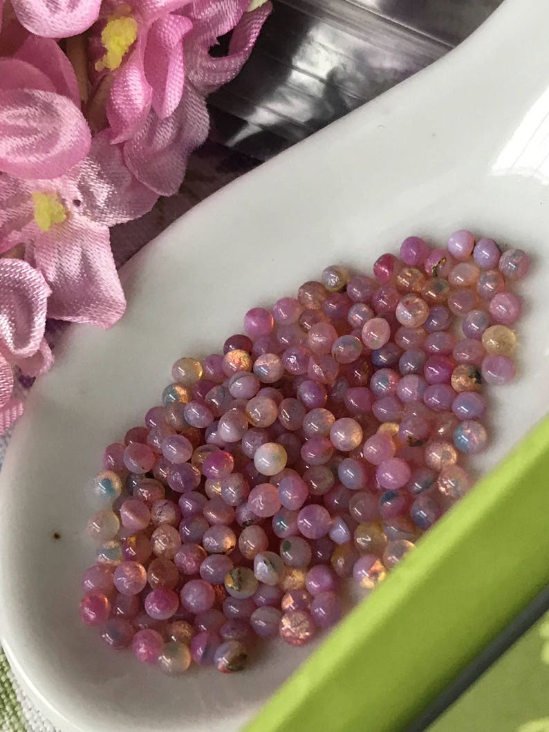 K&ouml;nnte beinhalten: Nahaufnahme einer wei&szlig;en L&ouml;ffel, gef&uuml;llt mit kleinen, runden, regenbogenfarbenen rosa Opalperlen.