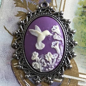May include: A purple cameo pendant with a silver filigree frame. The cameo features a white hummingbird in flight, surrounded by white flowers.