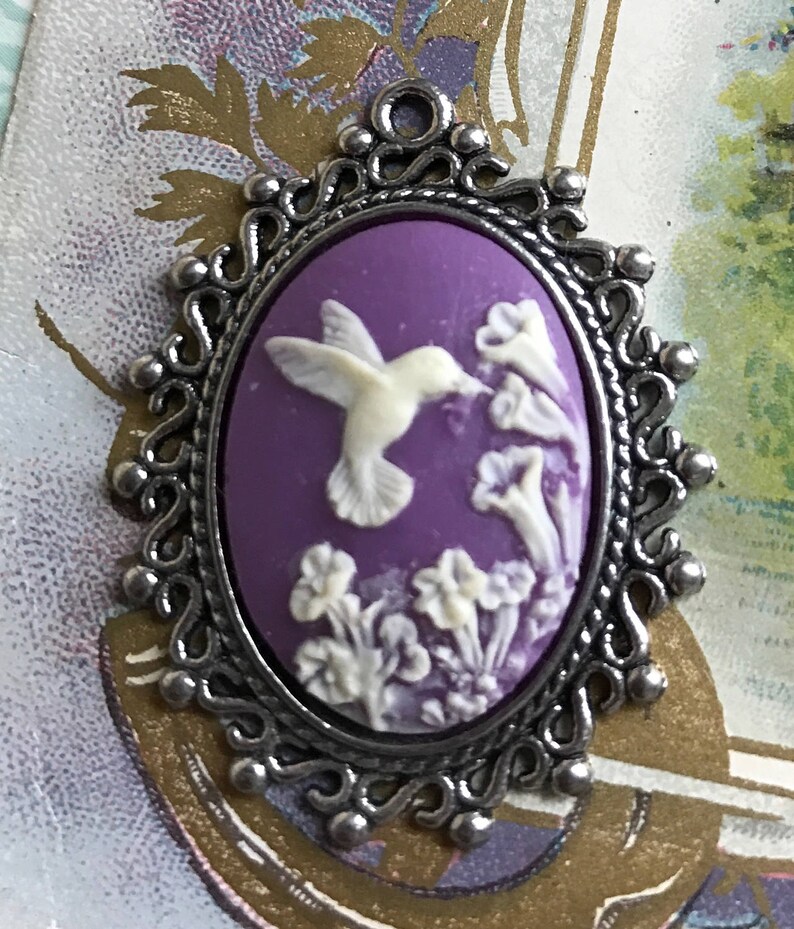 May include: A purple cameo pendant with a silver filigree frame. The cameo features a white hummingbird in flight, surrounded by white flowers.