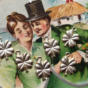 May include: Six silver four-leaf clover charms with a loop for hanging. The charms are on a green and white background with a vintage postcard image of a couple.