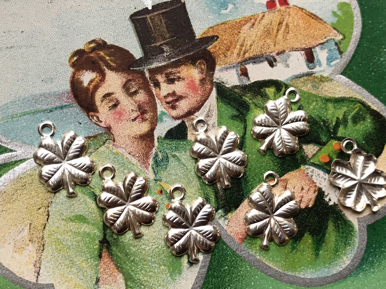 May include: Six silver four-leaf clover charms with a loop for hanging. The charms are on a green and white background with a vintage postcard image of a couple.