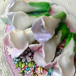 May include: A collection of porcelain flower embellishments in shades of white, green, and lavender. The flowers have a delicate, vintage aesthetic and are perfect for adding a touch of elegance to crafts and home decor.