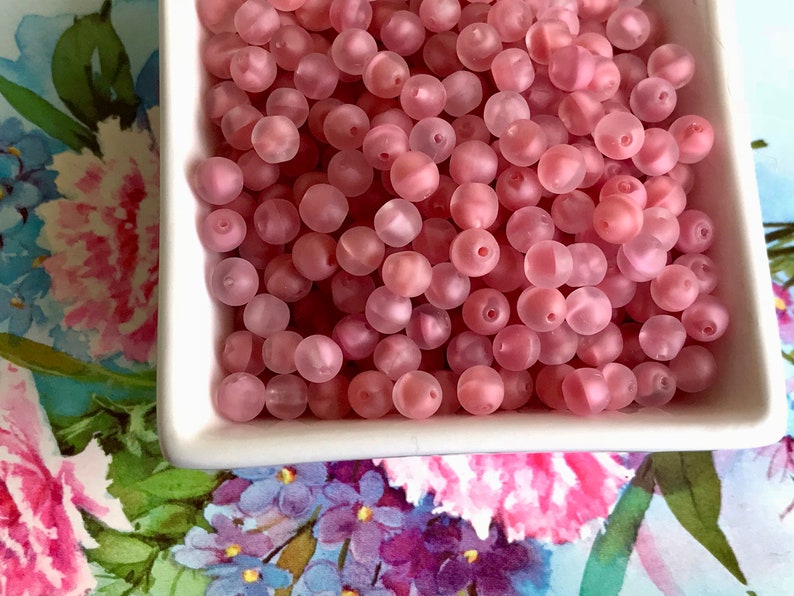 May include: A bowl filled with pink frosted glass beads. The beads are round and have a matte finish.