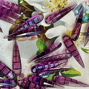 May include: A collection of purple glass teardrop-shaped beads with a faceted design. The beads are scattered on a floral patterned surface.