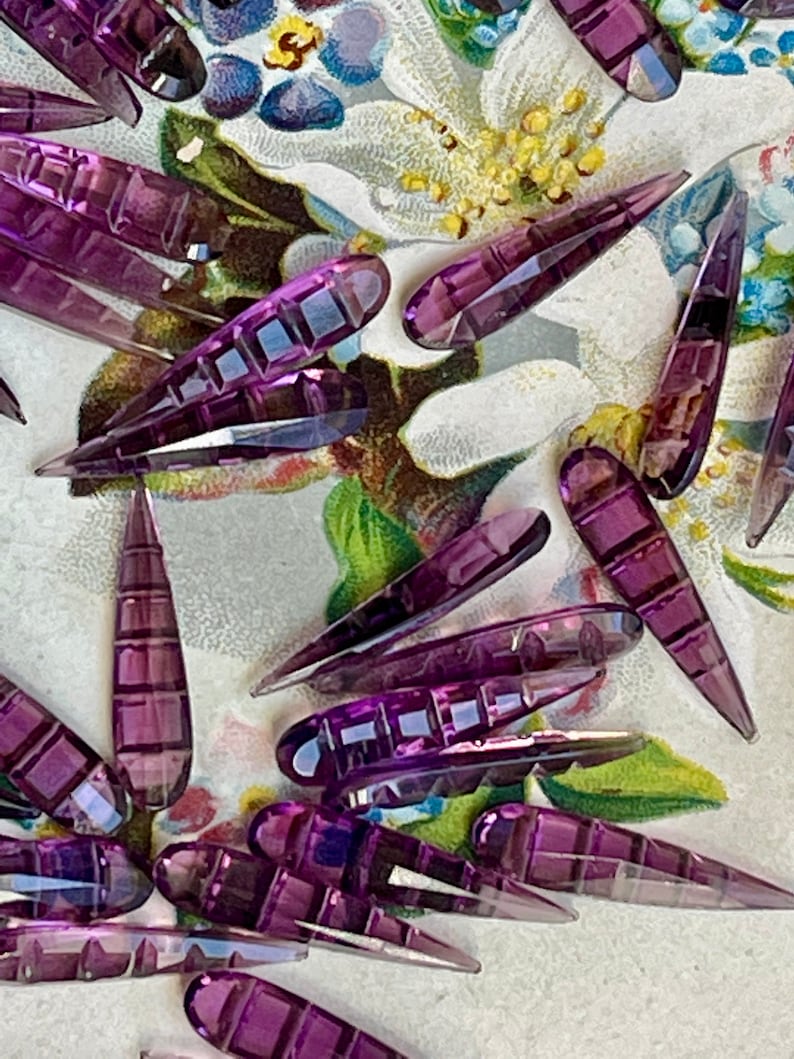 May include: A collection of purple glass teardrop-shaped beads with a faceted design. The beads are scattered on a floral patterned surface.