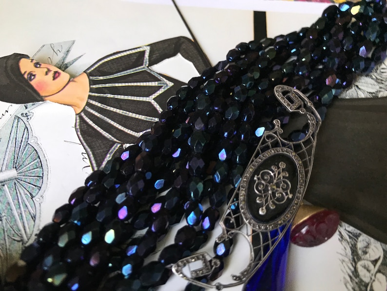 May include: A multi-strand necklace made of dark blue iridescent glass beads with a silver filigree clasp. The clasp features a black enamel oval with a silver floral design.