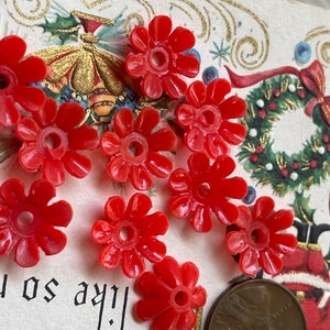 May include: Nine red plastic flower-shaped beads with a hole in the center. The beads are scattered on a white surface with a vintage Christmas card in the background.