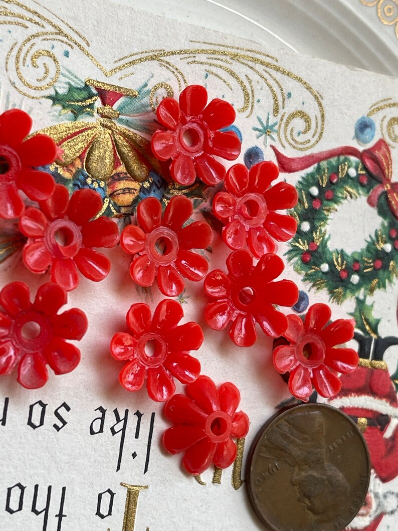 May include: Nine red plastic flower-shaped beads with a hole in the center. The beads are scattered on a white surface with a vintage Christmas card in the background.