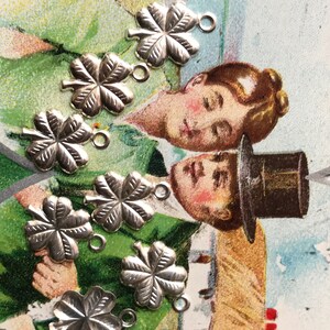 May include: Eight silver four-leaf clover charms with jump rings. The charms are on a vintage postcard with a green background.