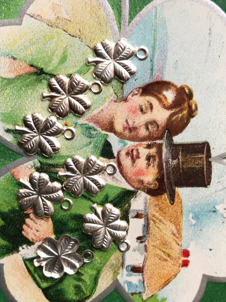 May include: Eight silver four-leaf clover charms with jump rings. The charms are on a vintage postcard with a green background.