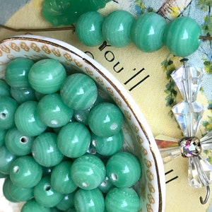 May include: A bowl filled with green glass beads. The beads have a swirl pattern and are smooth and shiny. The bowl is white with a gold rim.