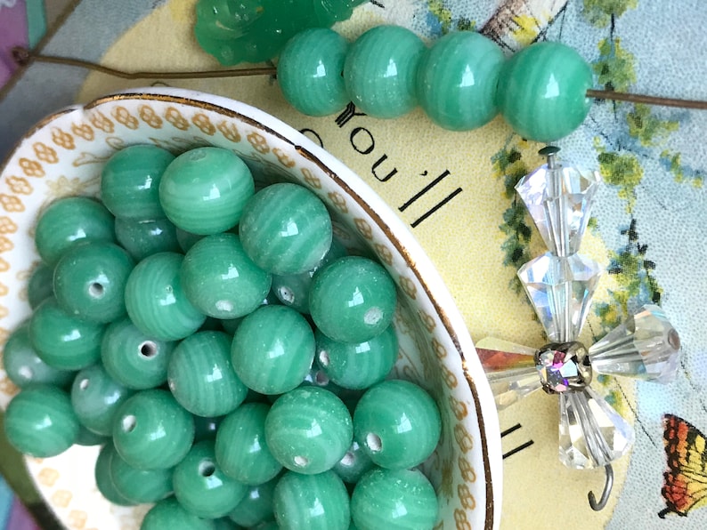 May include: A bowl filled with green glass beads. The beads have a swirl pattern and are smooth and shiny. The bowl is white with a gold rim.