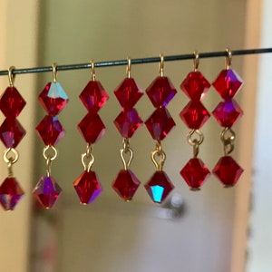 May include: A collection of red crystal earrings hanging from a wire. Each earring features three faceted, diamond-shaped crystals connected by gold-colored metal loops. The crystals have a vibrant, reflective quality.