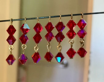 May include: A collection of red crystal earrings hanging from a wire. Each earring features three faceted, diamond-shaped crystals connected by gold-colored metal loops. The crystals have a vibrant, reflective quality.