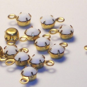 May include: A group of white glass rhinestones with gold metal settings and loops. The rhinestones are scattered on a white surface.