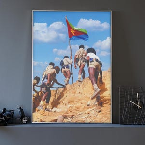 Könnte beinhalten: Ein gerahmter Druck zeigt eine Gruppe von Personen, die einen felsigen Hügel hinaufsteigen, wobei eine Person eine Flagge mit roten, grünen, gelben und blauen Farben vor blauem Himmel hält. Das Bild wird an einer grauen Wand angezeigt.