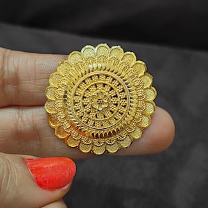 May include: A gold-colored ring with an intricate floral design. The ring features a central flower surrounded by detailed petal-like elements. The ring is held up by a person's fingers, with a dark background.