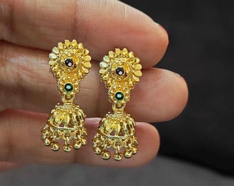 May include: A pair of gold-colored earrings with a floral design. Each earring features a flower-shaped top with a black and red center stone, a green stone, and a bell-shaped bottom with dangling gold beads. The earrings are held in a hand.