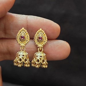 May include: A pair of gold earrings with a floral design. Each earring has a small, round, red gemstone at the centre of the top section. The bottom section is bell-shaped with intricate filigree detailing.