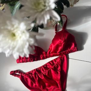 Könnte beinhalten: Ein rotes Satin-Dessous-Set, bestehend aus einem Triangel-BH und passendem Slip. Das Set wird auf einer weißen Oberfläche präsentiert, mit weißen Blumen im Hintergrund. Die Dessous haben zarte Träger und eine glatte, glänzende Textur.
