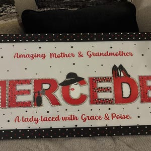 May include: A framed artwork with the name "MERCEDES" in large red letters, embellished with rhinestones. The artwork includes a black hat, high heels, and the text "Amazing Mother & Grandmother" and "A lady laced with Grace & Poise."