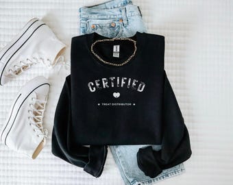 May include: A black sweatshirt with the word "CERTIFIED" in white, a heart graphic, and the text "TREAT DISTRIBUTOR". The sweatshirt is paired with light-wash jeans, a gold chain, and white high-top sneakers.