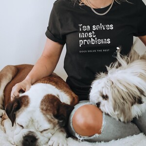 Op de afbeelding: Een vrouw draagt een zwart T-shirt met de tekst "Tea solves most problems. Dogs solve the rest."  Ze zit met twee honden, een bruin-witte en een witte.