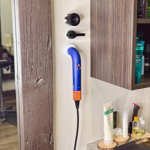 May include: A blue and orange hair dryer with a long cord is mounted on a white wall. The hair dryer is attached to a white wall mount. The hair dryer is in a salon setting.